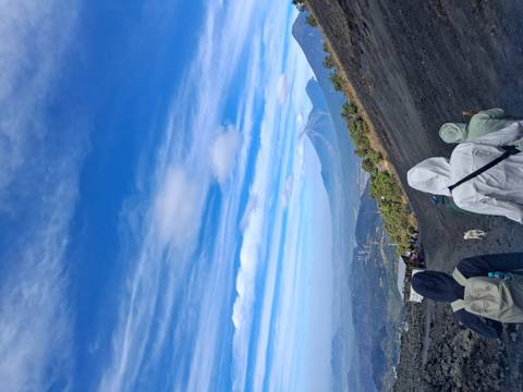       Hikers ascend the dark volcanic slope of Pacaya with plume-capped volcano in view.
  