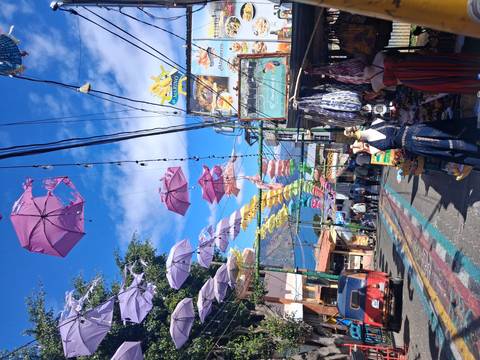       Vibrant street market with colorful umbrellas strung overhead against a bright sky.
  