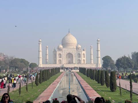       Central perspective of the Taj Mahal with long water channel and crowds lining the gardens under hazy blue sky
  