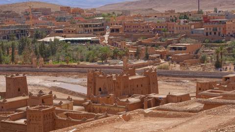       Panoramic view of the earthen kasbahs and palm groves of Ait Benhaddou set against arid hills.
  