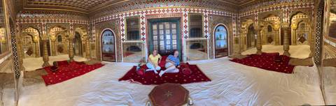       Two travellers relax on cushions in a lavish Rajasthani palace room adorned with mirrors and red carpets.
  