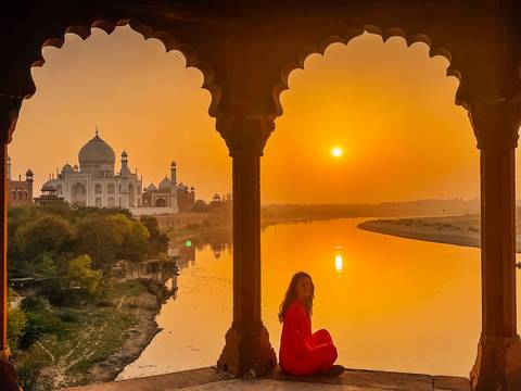       Woman in red looks across the Yamuna River to the Taj Mahal at sunrise, framed by ornate arch columns
  