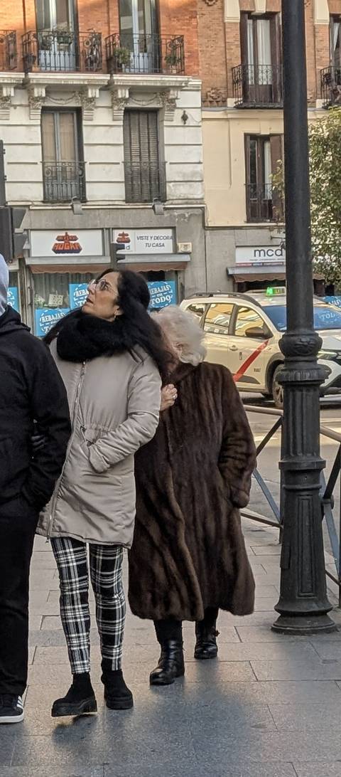      Older and younger women bundled in coats wait at a city crossing.
  