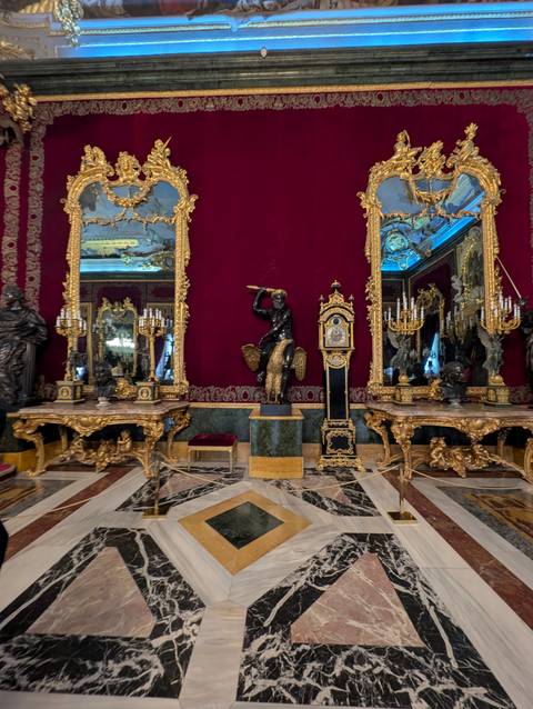       Lavish red-and-gold palace room filled with ornate clocks, mirrors and candelabra.
  
