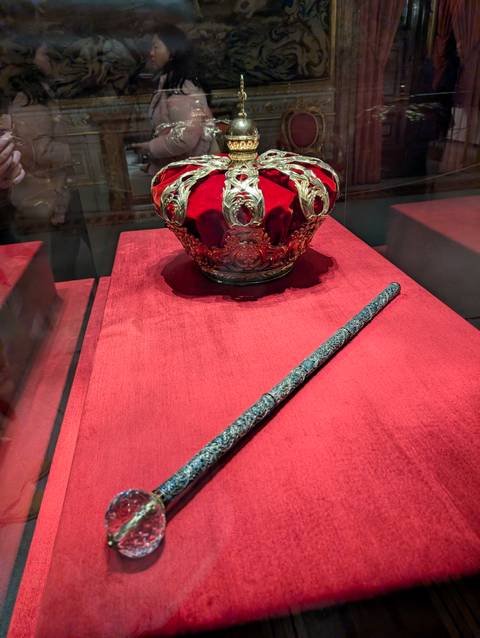       Regal crown and ornate sceptre displayed on rich red velvet inside a museum case.
  