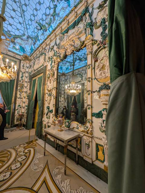       Opulent mirror-lined salon with chandeliers, marble floors and ornate green drapery.
  