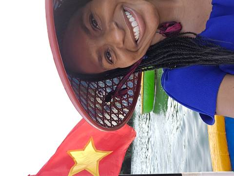       Close-up selfie of a smiling traveler wearing a Vietnamese hat on a boat with the national flag behind.
  