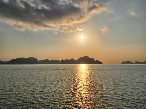       The sun sets over limestone islands of Halong Bay casting a golden reflection on calm water.
  