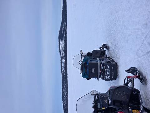       Two snowmobiles parked on a vast frozen lake with forested hills in the distance under a gray sky.
  