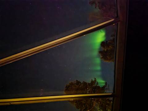       Northern lights seen through the angled glass window of a cabin among silhouetted trees at night.
  
