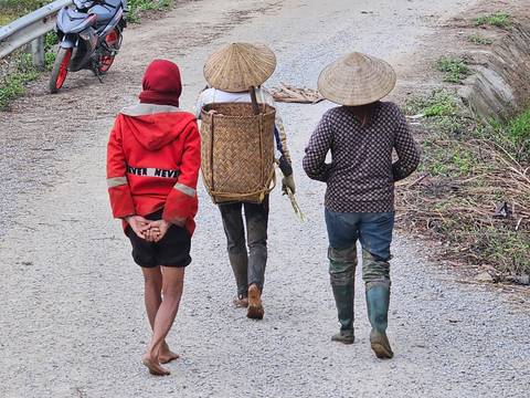       Three local villagers walk along a rural gravel road, one carrying a woven basket backpack
  