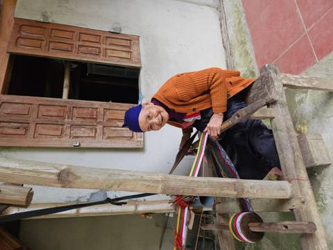       Elderly woman smiling while weaving colourful fabric on a traditional loom in a village house
  