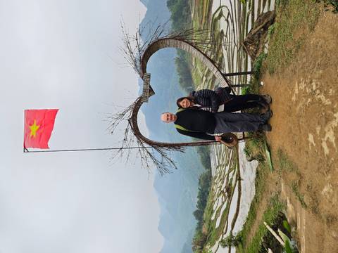       Travelling couple pose under a heart-shaped wooden arch with terraced hills and mountains behind
  