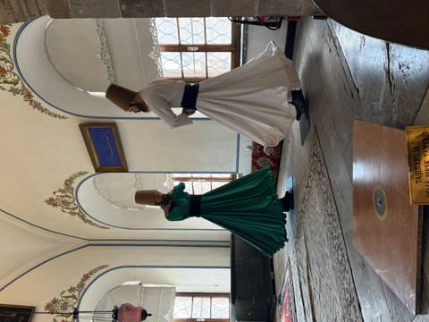       Two whirling dervishes in green and white robes perform inside a historic hall.
  