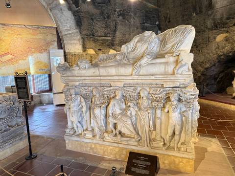       Marble sarcophagus with detailed reliefs displayed in a small museum room.
  
