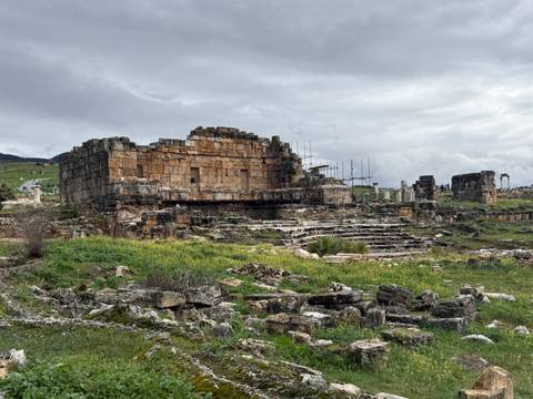       Partially restored ancient stone structure with scaffolding amid grassy ruins.
  