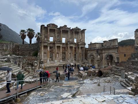       The grand facade of the Library of Celsus with visitors walking under umbrellas.
  
