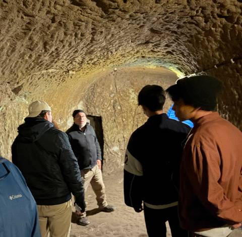       Tour guide speaks to visitors inside a dimly lit underground cave passage.
  