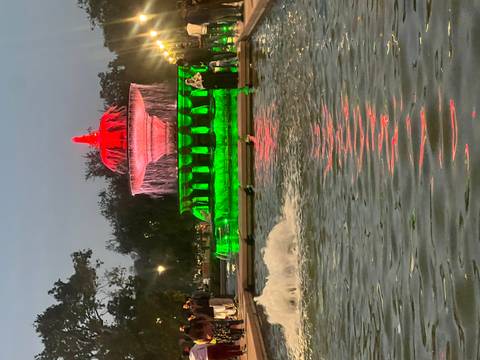       Nighttime fountain near India Gate lit in red and green with onlookers by the reflective pool.
  