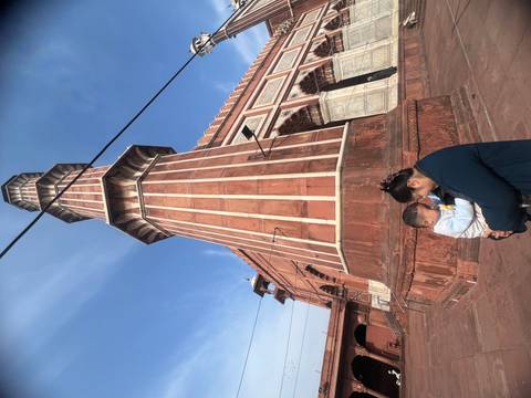       Mother holding a small child at the base of a red-sandstone mosque minaret under a bright blue sky.
  