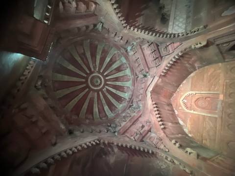       Dim interior view of a carved stone dome ceiling with radial floral patterns inside an Indian monument.
  