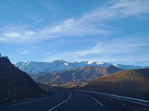       Snow-capped Atlas Mountains rising above rolling brown foothills and a curving highway at dawn light.
  