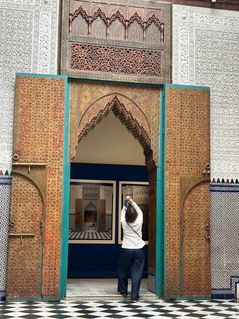       Tourist photographing an intricately tiled and carved gateway with vivid turquoise accents in Fes.
  