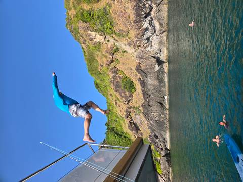       Man mid-air jumping off a boat into emerald sea with companions swimming below near rocky Thai coast.
  