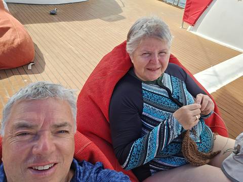       Selfie of two cruisers relaxing on deck chairs; woman knitting while man smiles at camera.
  