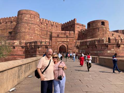       Older couple posing on the bridge leading to Agra Fort with red sandstone walls and crowds behind them
  