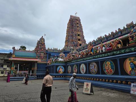       Vibrantly painted Hindu temple tower and ornately decorated walls with a few visitors in the courtyard.
  