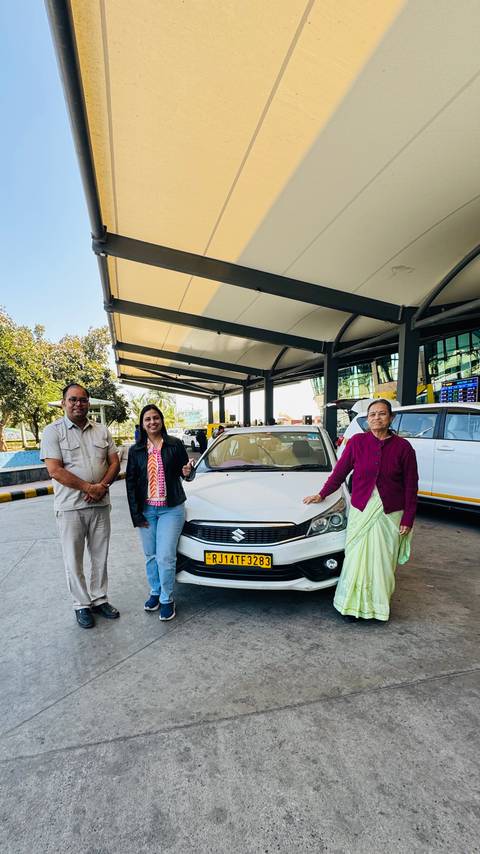       Tour guide, female traveller and elder woman posing in front of white Suzuki taxi outside Delhi airport
  