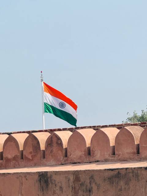       Indian national flag waving atop a historic sandstone wall against a pale sky.
  