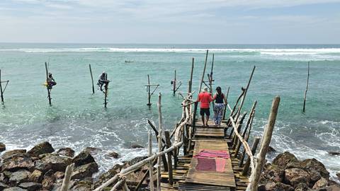       Tourists stand on wooden jetty watching traditional stilt fishermen over teal ocean waves.
  