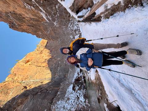       Couple with trekking poles stand on a snowy trail beside a stream with towering red cliffs in Zion.
  