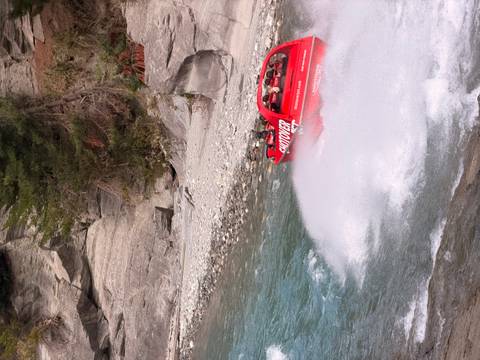       Red Shotover jet boat speeds through turquoise canyon waters leaving a trail of spray.
  