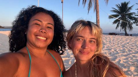       Two female friends smiling for a selfie on a white-sand beach lit by warm golden-hour light, with palm tree in background
  