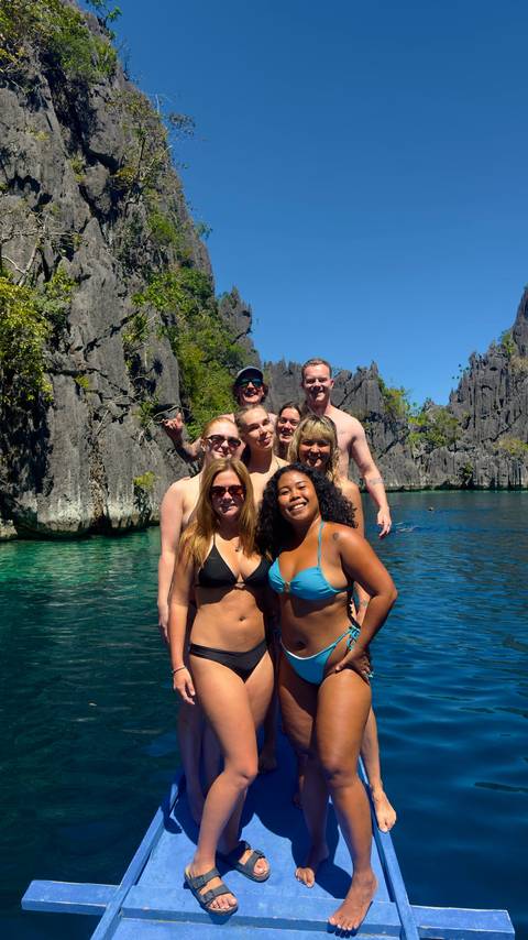       Group of travellers posing together in bikinis and swim trunks in a crystal-clear lagoon framed by limestone cliffs
  