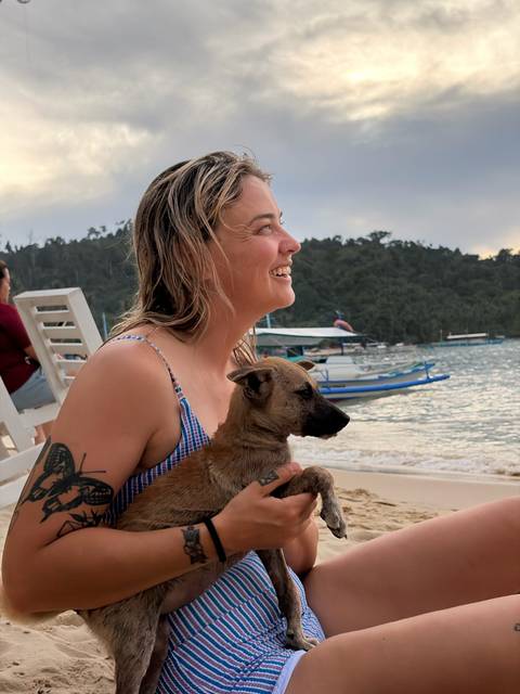       Smiling woman sitting on beach gently holding a small brown puppy with boats and forested hills in background
  
