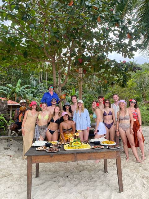       Large group of swimsuit-clad travellers posing under tropical trees on a beach holding coconuts
  