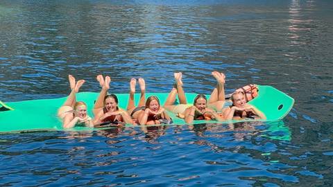       Five friends lying on a floating aqua mat, feet up, laughing on clear blue water
  