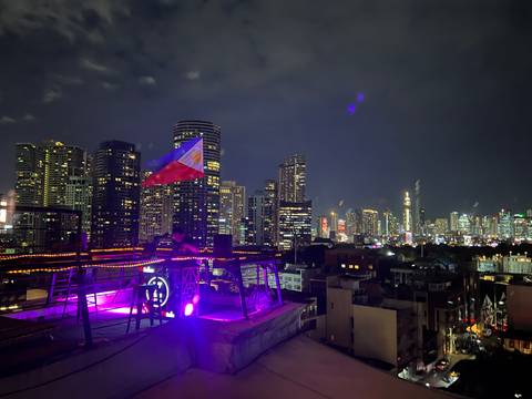       Nighttime skyline of Manila with brightly lit modern skyscrapers and a Philippine flag projection seen from a rooftop bar
  