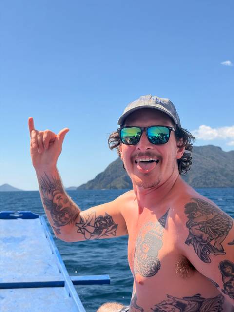       Man on boat flashes shaka sign with tongue out, wearing mirrored sunglasses against island backdrop.
  