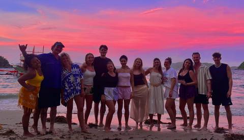       Group of travellers poses on beach with vivid pink and orange sunset sky behind.
  
