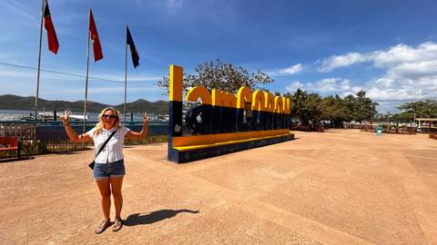       Traveler raises arms beside giant yellow 'I am Coron' sign near the waterfront on sunny day.
  