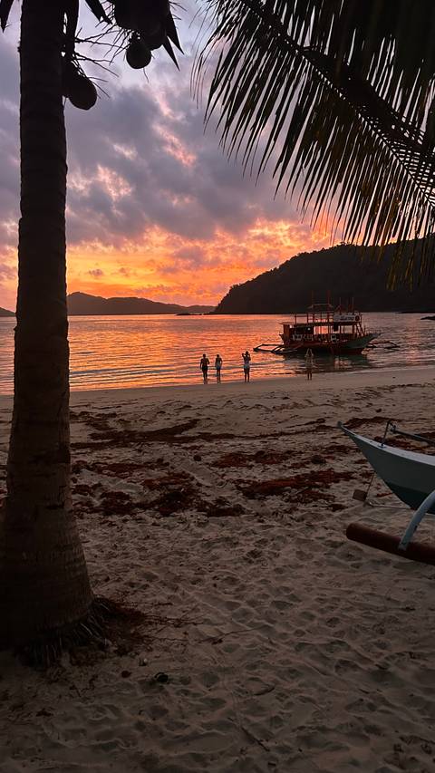       Small group standing ankle-deep in water watching vibrant orange sunset with traditional boat nearby.
  