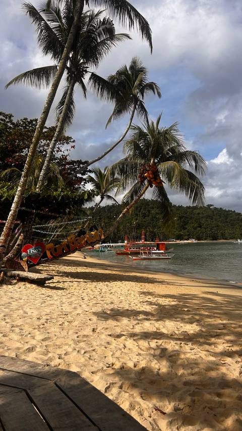       'Port Barton' beach sign hangs from palm beside anchored orange boats and jungle shoreline.
  