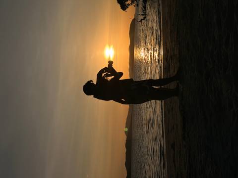       Silhouetted couple embracing while taking a photo at golden sunset on a quiet beach with calm water and distant islands.
  