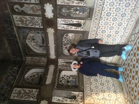       Couple wearing protective shoe covers inside an ornately tiled Mughal chamber.
  