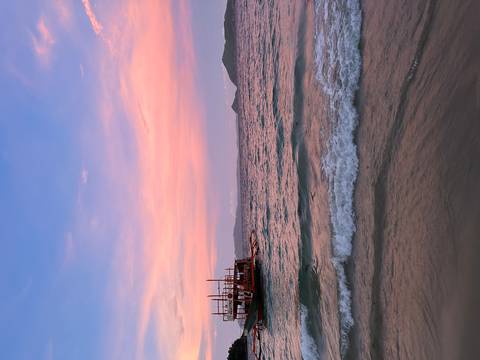       Pastel pink and orange sunset over gentle waves with a traditional wooden boat offshore.
  